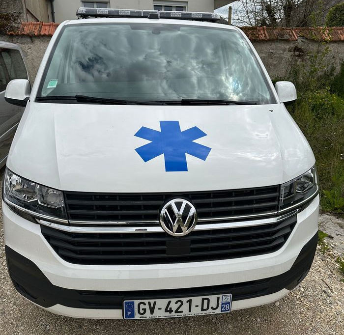 Ambulance Volkswagen blanche avec une étoile bleue, stationnée sur un chemin en gravier.