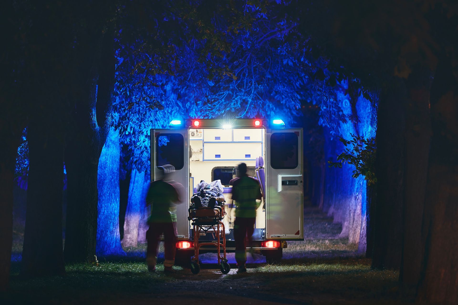 Ambulance avec portes arrière ouvertes, ambulanciers chargeant un brancard de nuit avec gyrophares bleus.