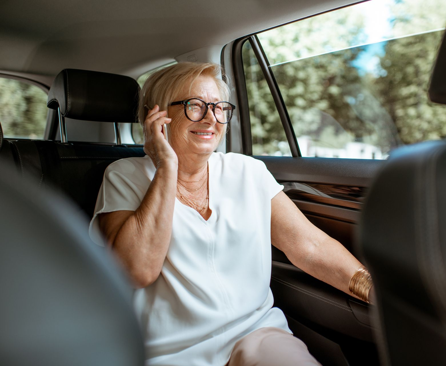 Femme en voiture, parlant au téléphone, souriante. Elle porte des lunettes et un haut blanc.