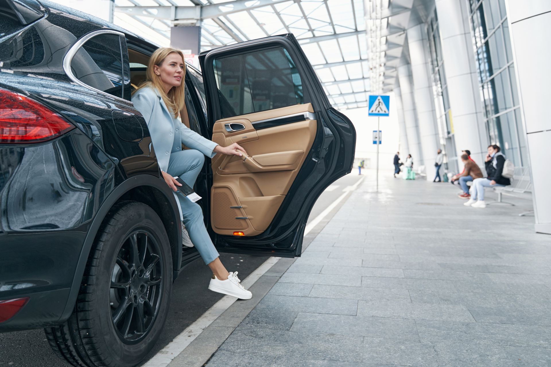 Une femme sort d'un SUV noir à l'aéroport. Elle porte un tailleur bleu et des baskets blanches.