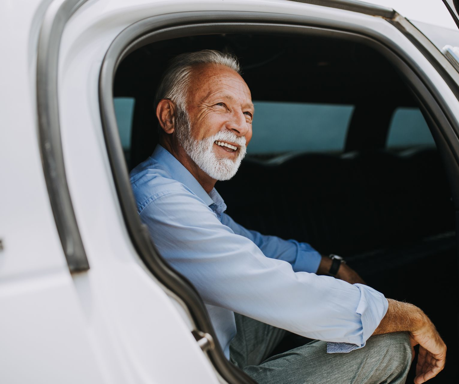 Un homme âgé souriant, assis dans une voiture, regarde sur le côté, la portière ouverte.