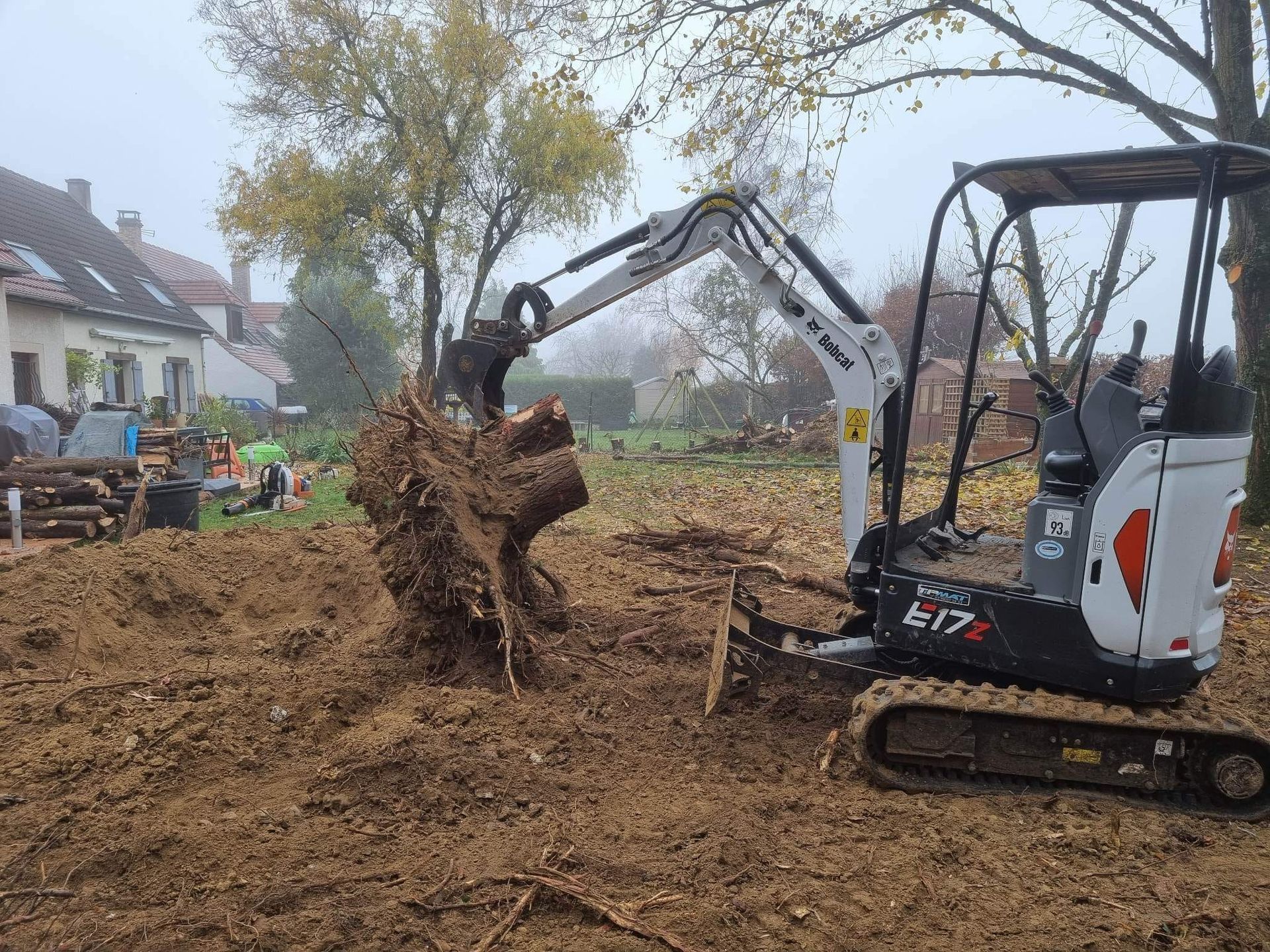 Une pelleteuse soulève une souche d'arbre dans une cour boueuse près des maisons par une journée brumeuse.