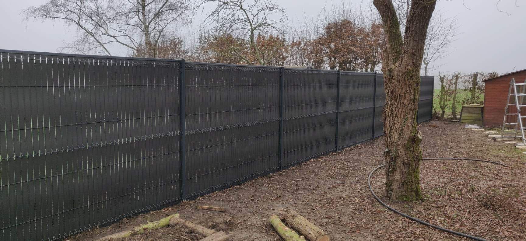 Une longue clôture noire préserve l'intimité, bordant des arbres dénudés et un petit abri de jardin dans une cour gravillonnée.