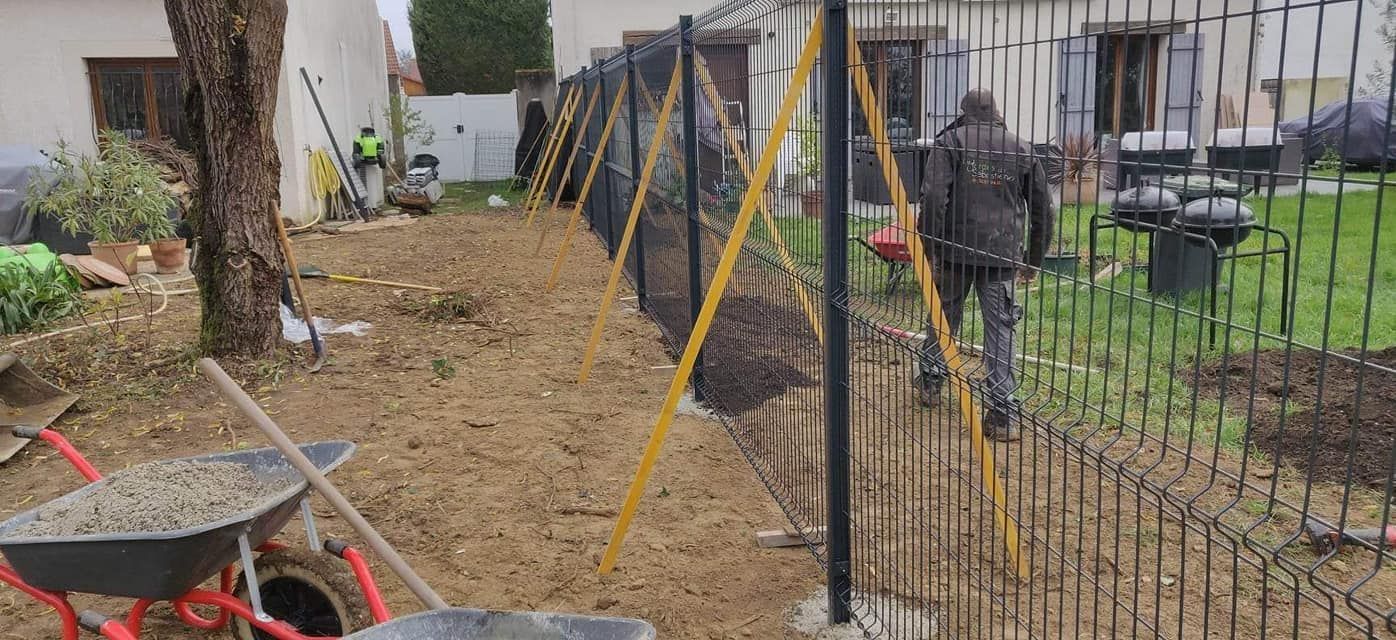 Un étroit chemin de terre longe un bâtiment et une clôture en fil de fer, avec des outils de jardin et une brouette dans une cour.