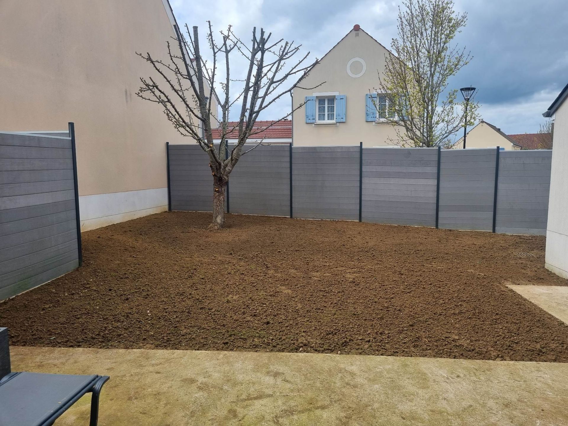 Petite cour arrière clôturée avec paillis brun, arbre sans feuilles, panneaux gris et maisons voisines visibles.