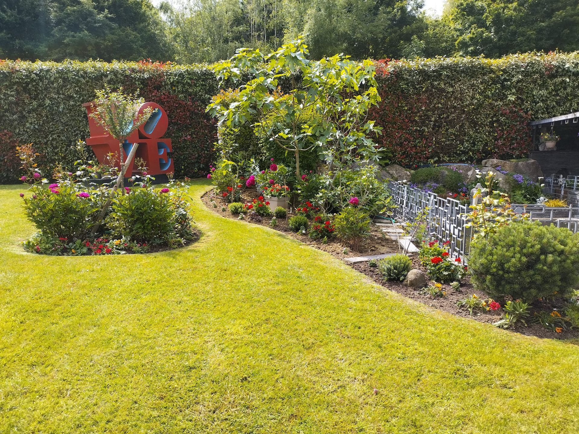 Jardin ensoleillé avec une pelouse verte, des fleurs colorées et une sculpture décorative près d'une haie.