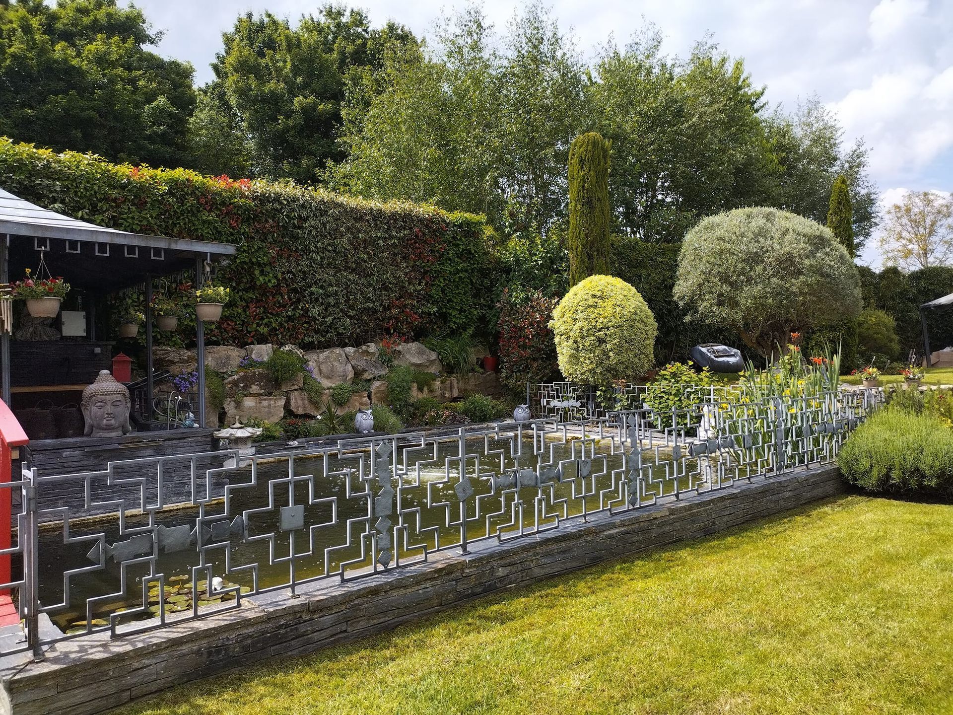 Jardin paysager avec haies taillées, clôture métallique et terrasse ombragée donnant sur une pelouse.