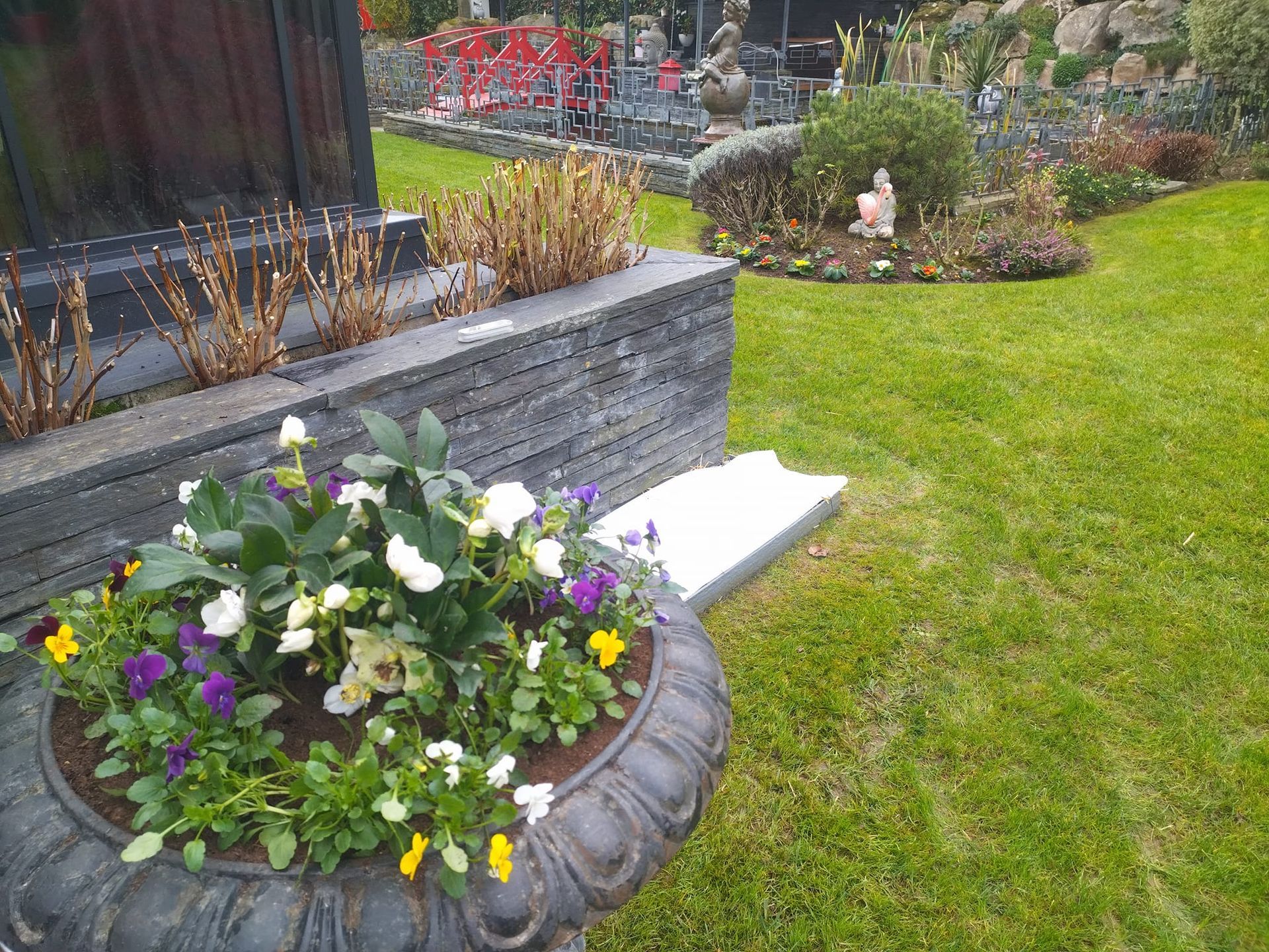Une jardinière remplie de fleurs à côté d'un parterre surélevé sur la pelouse, avec une petite figurine de jardin à proximité.
