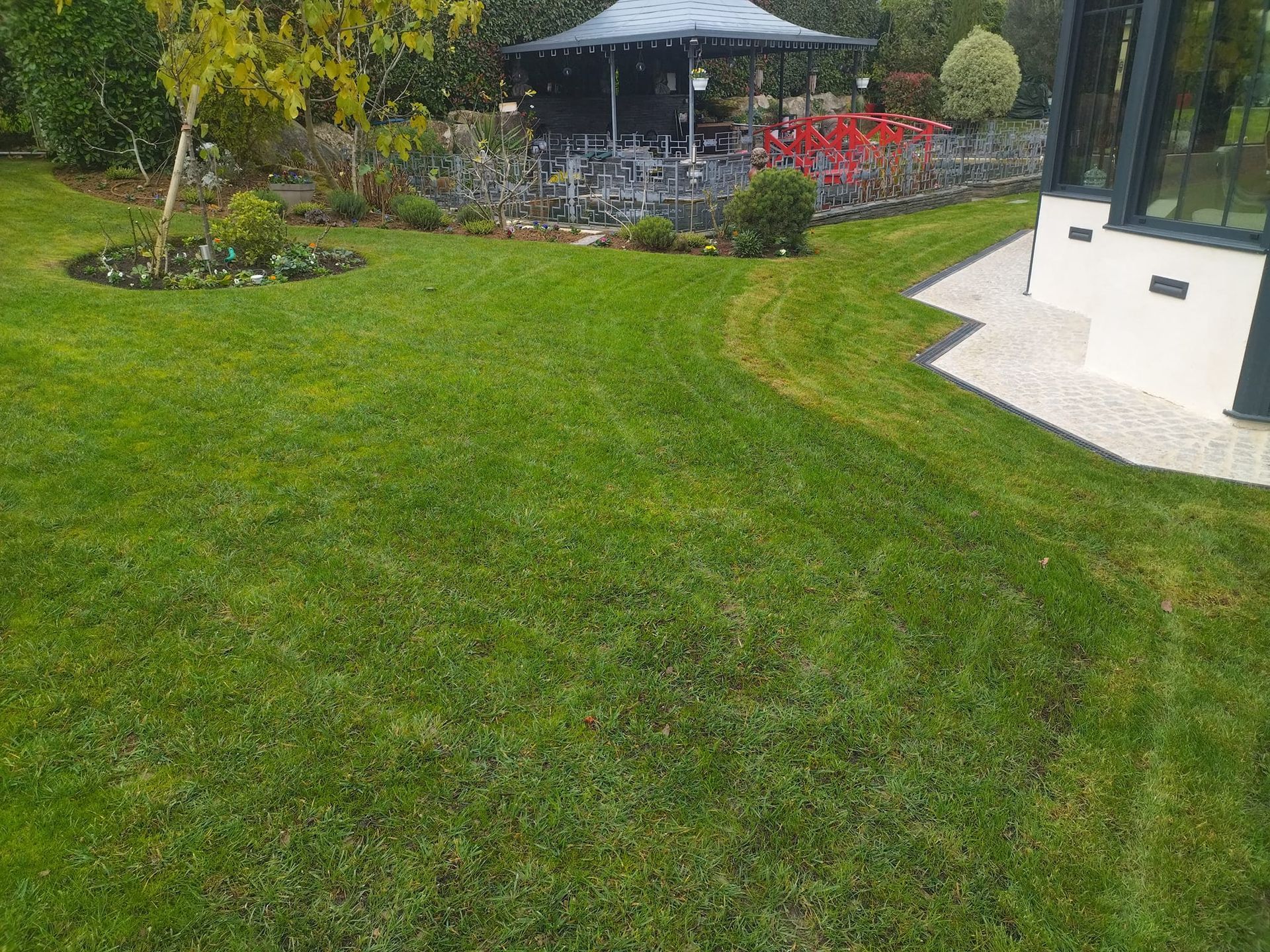 Pelouse verte avec un kiosque, des parterres de fleurs et un bâtiment blanc au bord d'un chemin sinueux.
