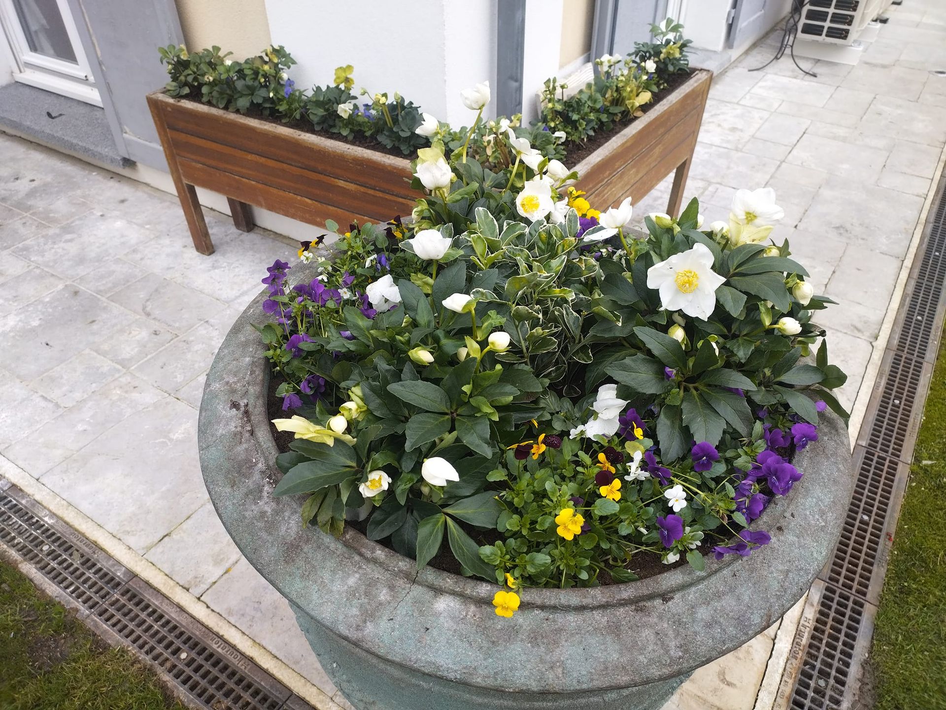 Jardinière ronde en pierre avec des fleurs blanches, jaunes et violettes à côté d'un banc sur un trottoir.