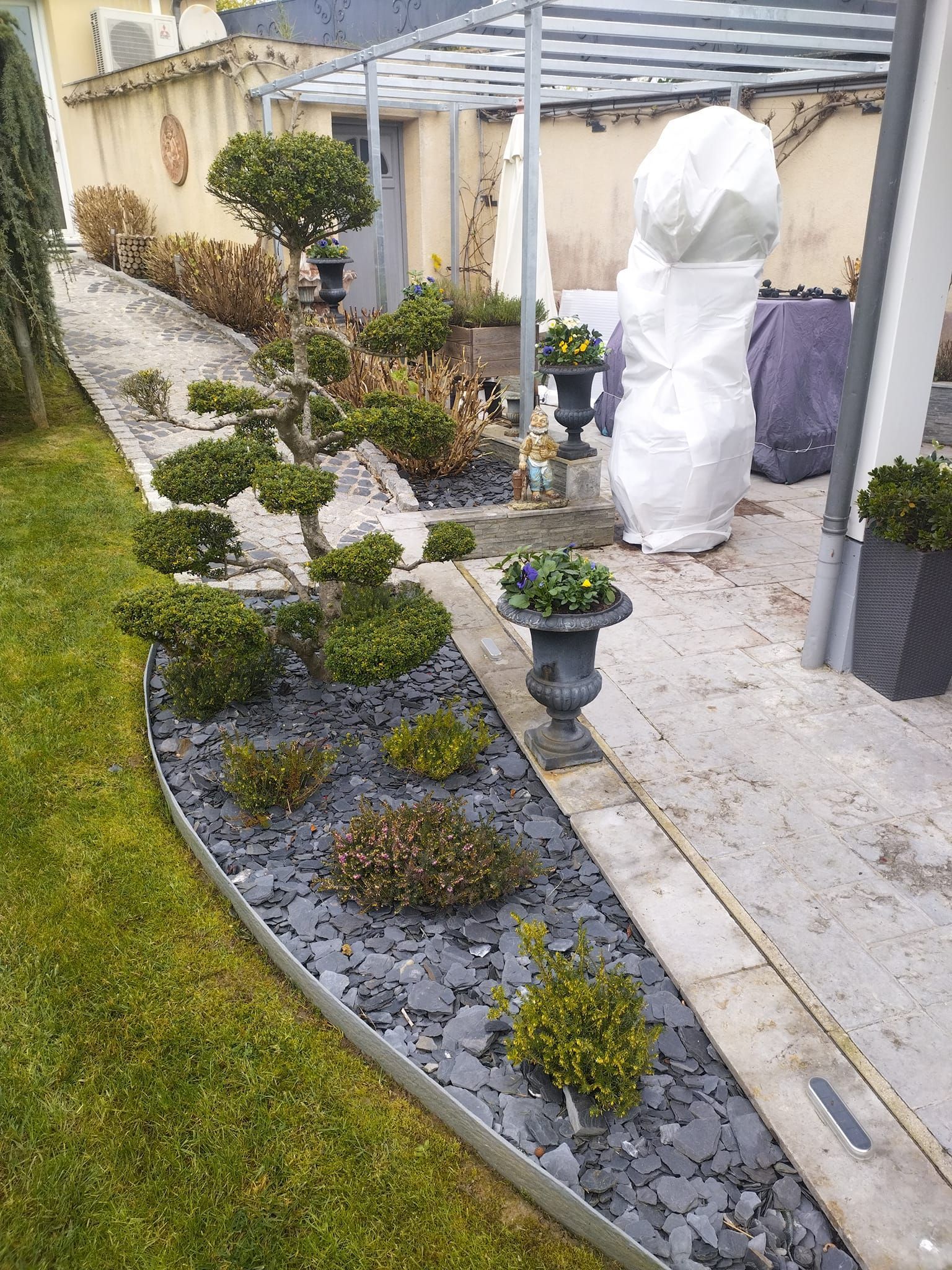 Allée de jardin à l'arrière d'une maison, avec plantes en pot, paillis foncé et objets recouverts.