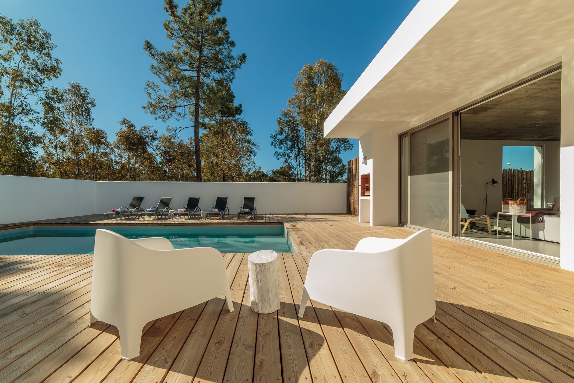 Patio moderne avec chaises blanches, terrasse en bois, piscine et maison aux parois de verre sous un ciel bleu azur.