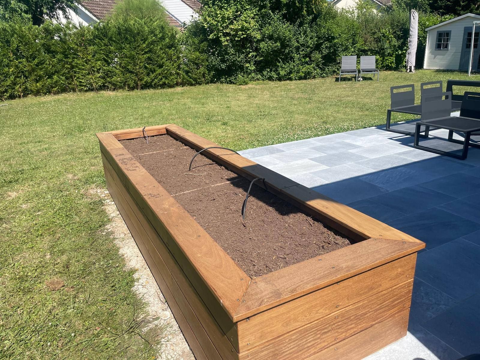 Jardinière surélevée en bois remplie de terre, à côté d'une terrasse dans un jardin gazonné.