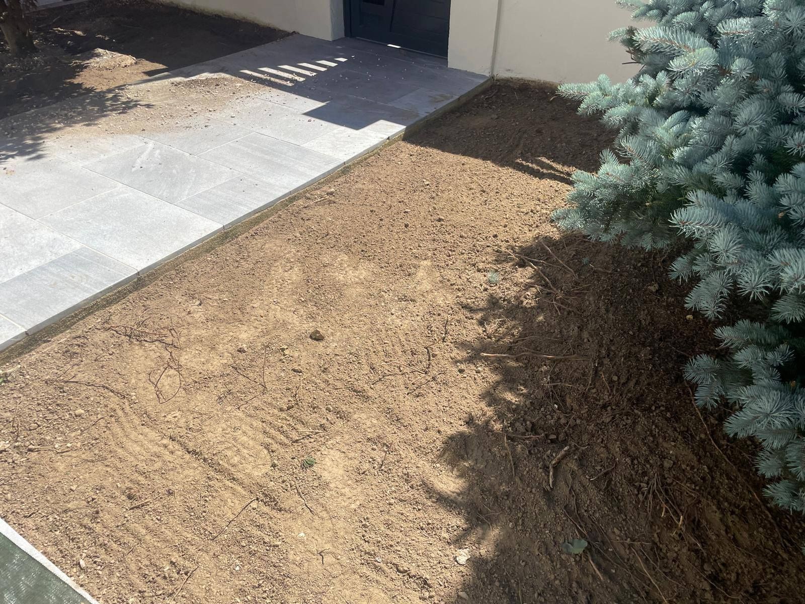 Massif de terre nue à côté d'une allée en béton et d'un conifère près de l'entrée d'une maison.