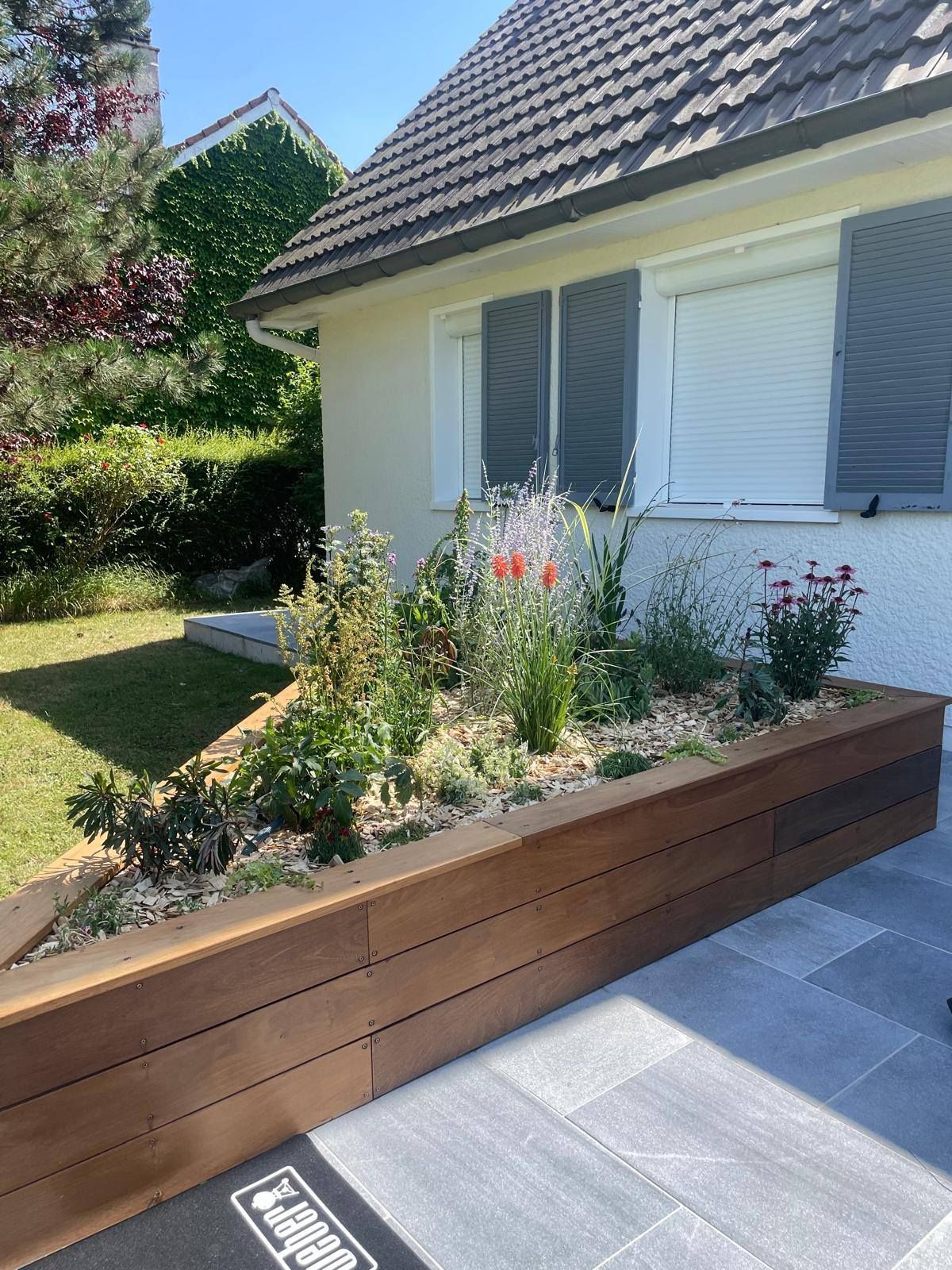 Jardinière surélevée en bois avec des fleurs à côté d'une maison blanche et de dalles de terrasse grises au soleil.