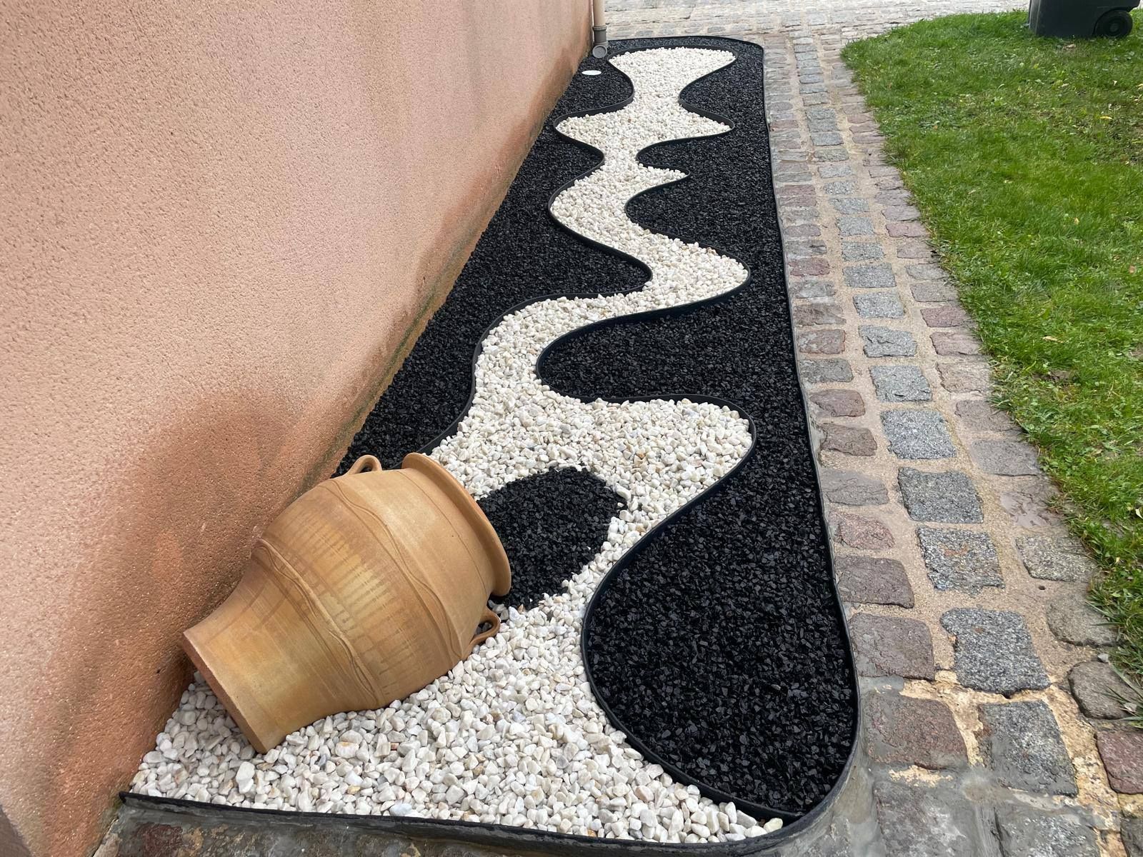 Allée de jardin décorative en pierres noires, blanches et grises, bordée d'un mur et de gazon, avec un tonneau en bois renversé pour la jardinière.