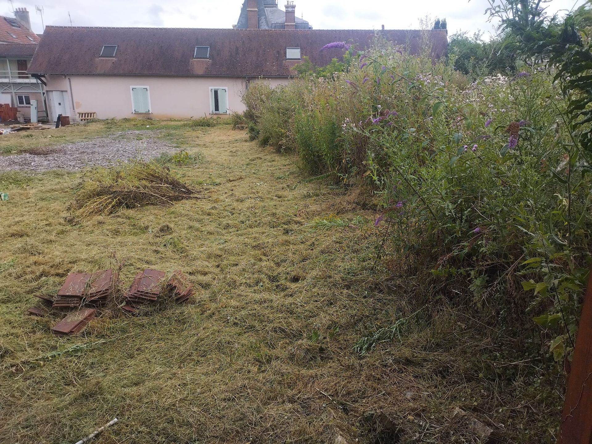 Cour arrière avec pelouse tondue, zone de gravier, arbustes et une maison rose avec des lucarnes.
