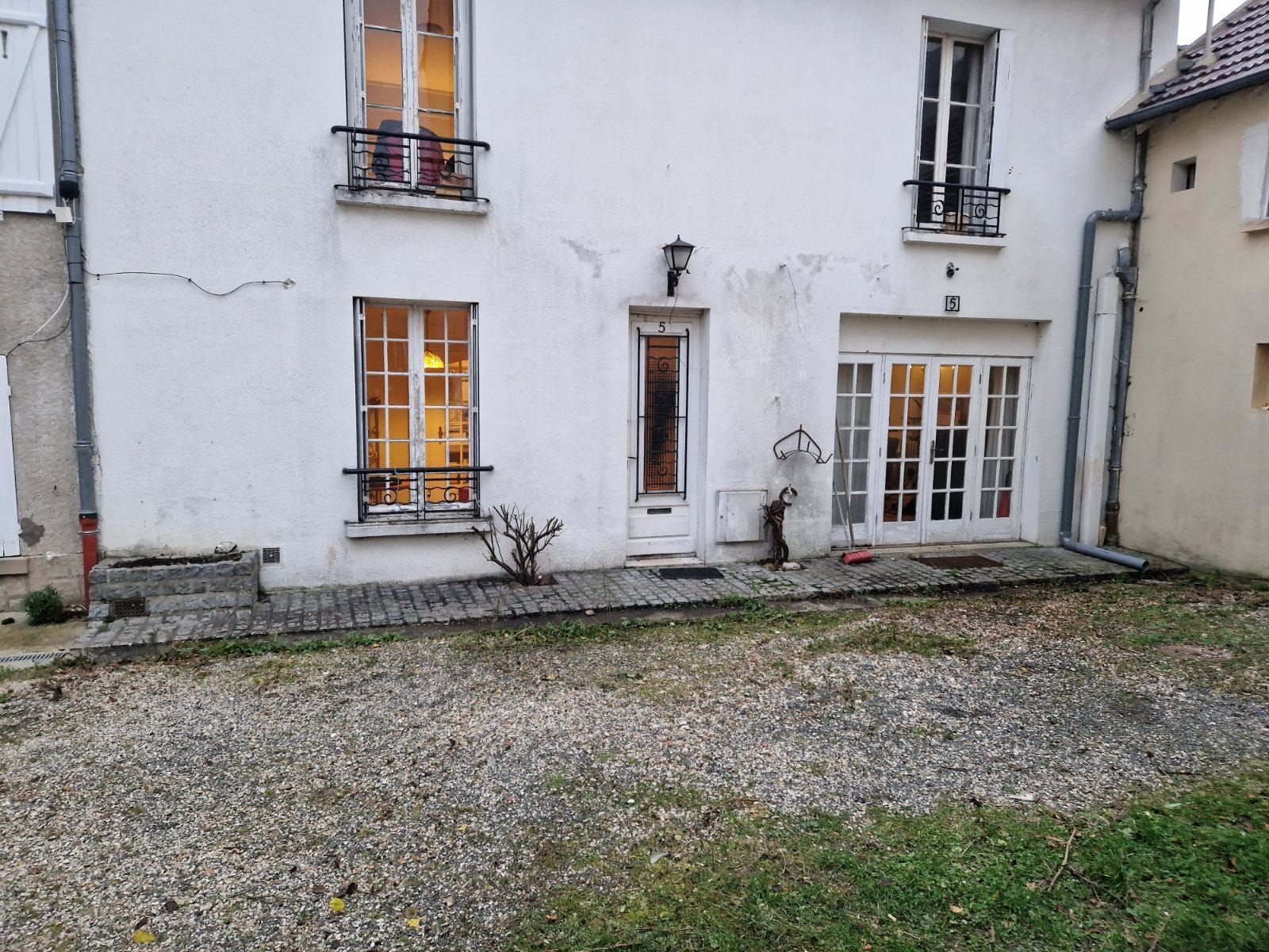 Façade de maison en pierre blanche avec fenêtres éclairées, porte d'entrée, petite statue et cour gravillonnée.