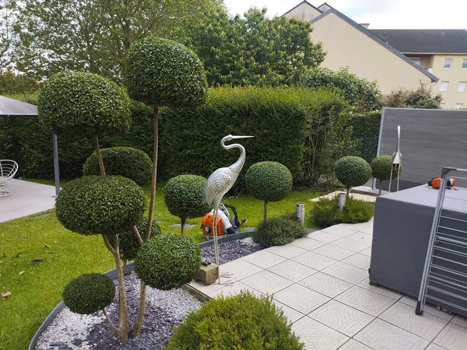 Jardin avec des topiaires taillées, une statue de grue blanche et une terrasse pavée jouxtant une maison moderne.