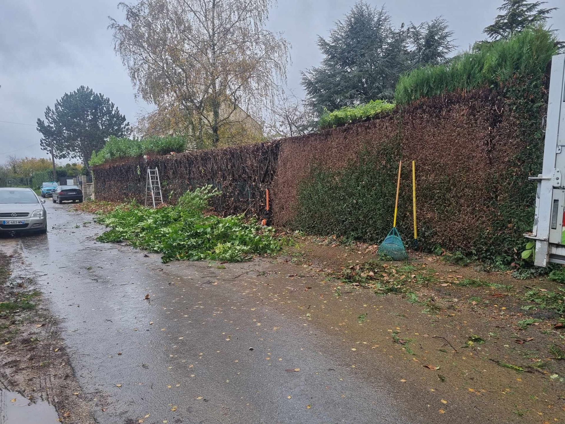 Rue résidentielle pluvieuse avec une voiture garée, une haie et un balai près d'un portail.