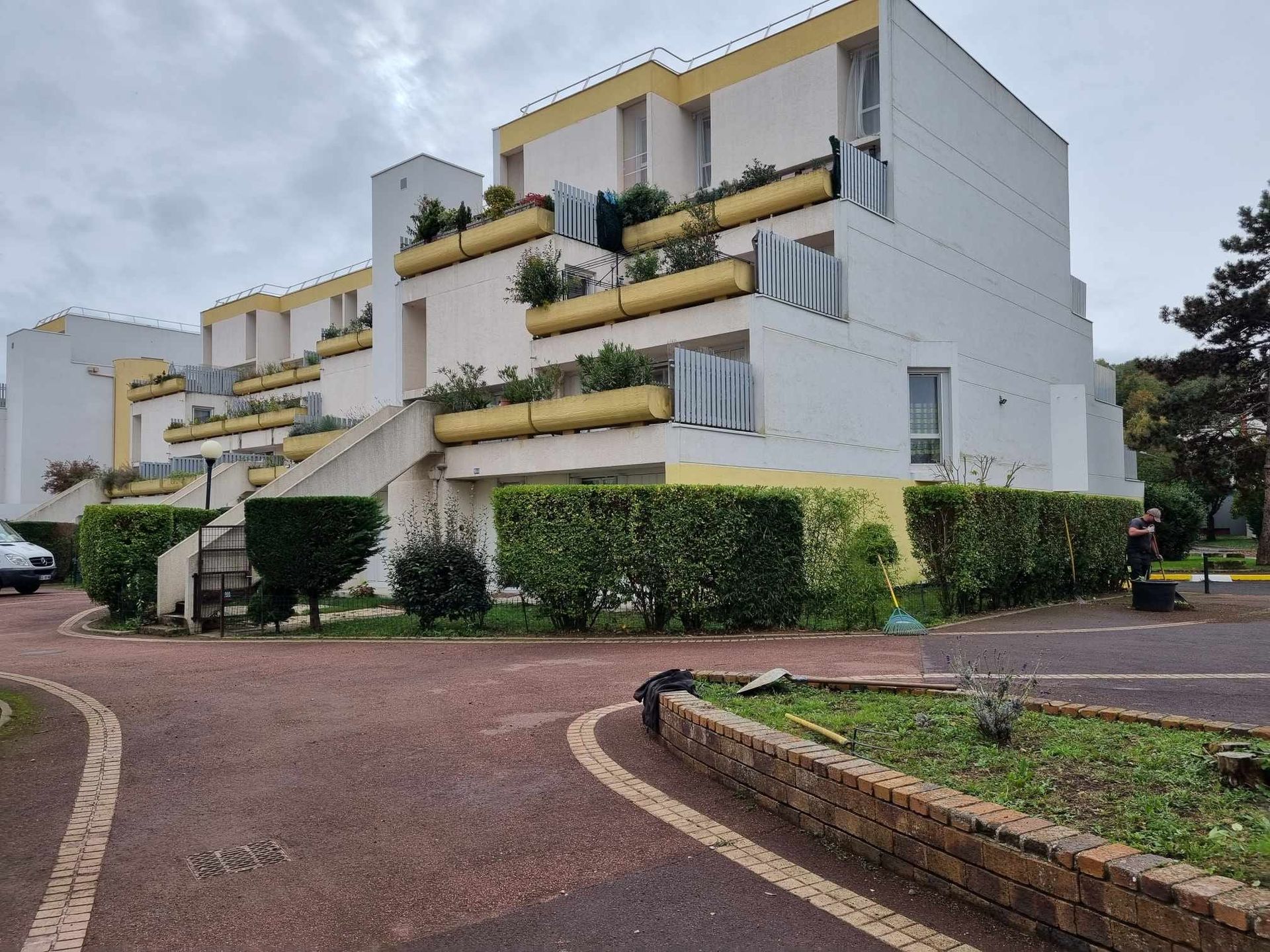 Immeuble d'appartements moderne blanc avec des touches de jaune et des balcons, donnant sur une allée pavée et un jardin bordé de haies.