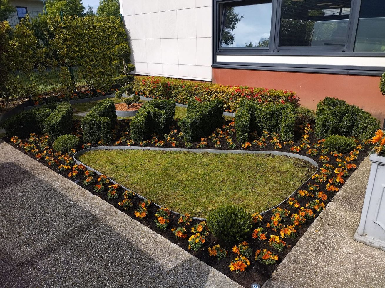 Petit parterre de jardin paysager avec des fleurs orange, des arbustes et une bordure de pierre incurvée, à côté d'un bâtiment.