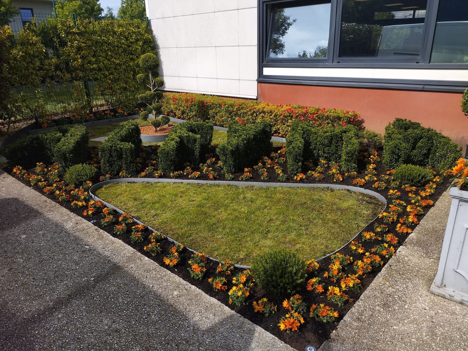Petit parterre de jardin paysager avec des fleurs orange, des arbustes et une bordure de pierre incurvée, à côté d'un bâtiment.