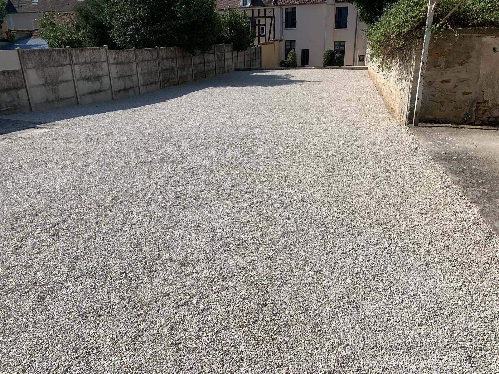 Allée de gravier menant à une maison, bordée de murs de pierre et de verdure baignée de soleil.