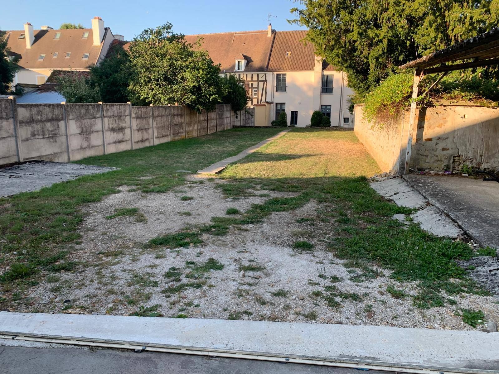 Allée de gravier vide menant à une maison, bordée de murs et d'arbres baignée par la douce lumière du soir.