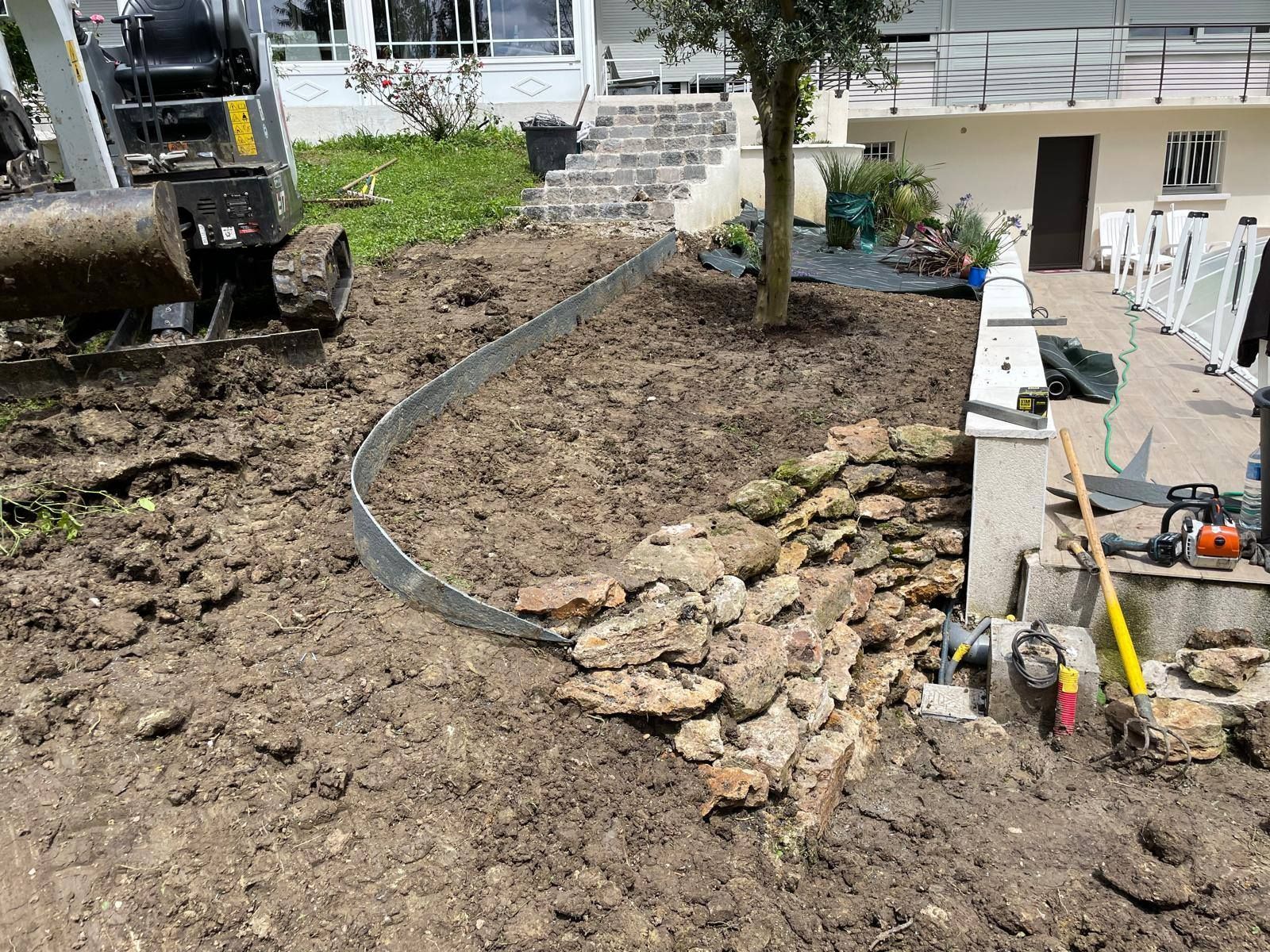 Travaux d'excavation dans un jardin près d'une maison, avec des tas de terre, des pierres et une pelle mécanique.