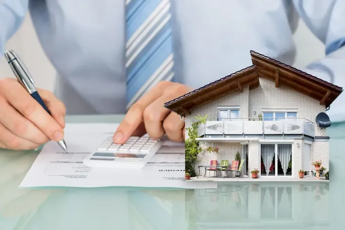 Un hombre está usando una calculadora y escribiendo en un trozo de papel al lado de una casa modelo.