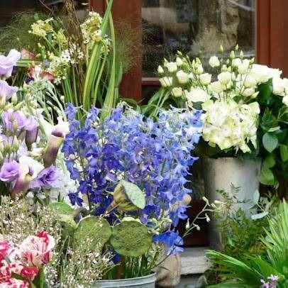 Des bouquets colorés de fleurs blanches, violettes, bleues et roses dans des pots devant la vitrine d’une boutique