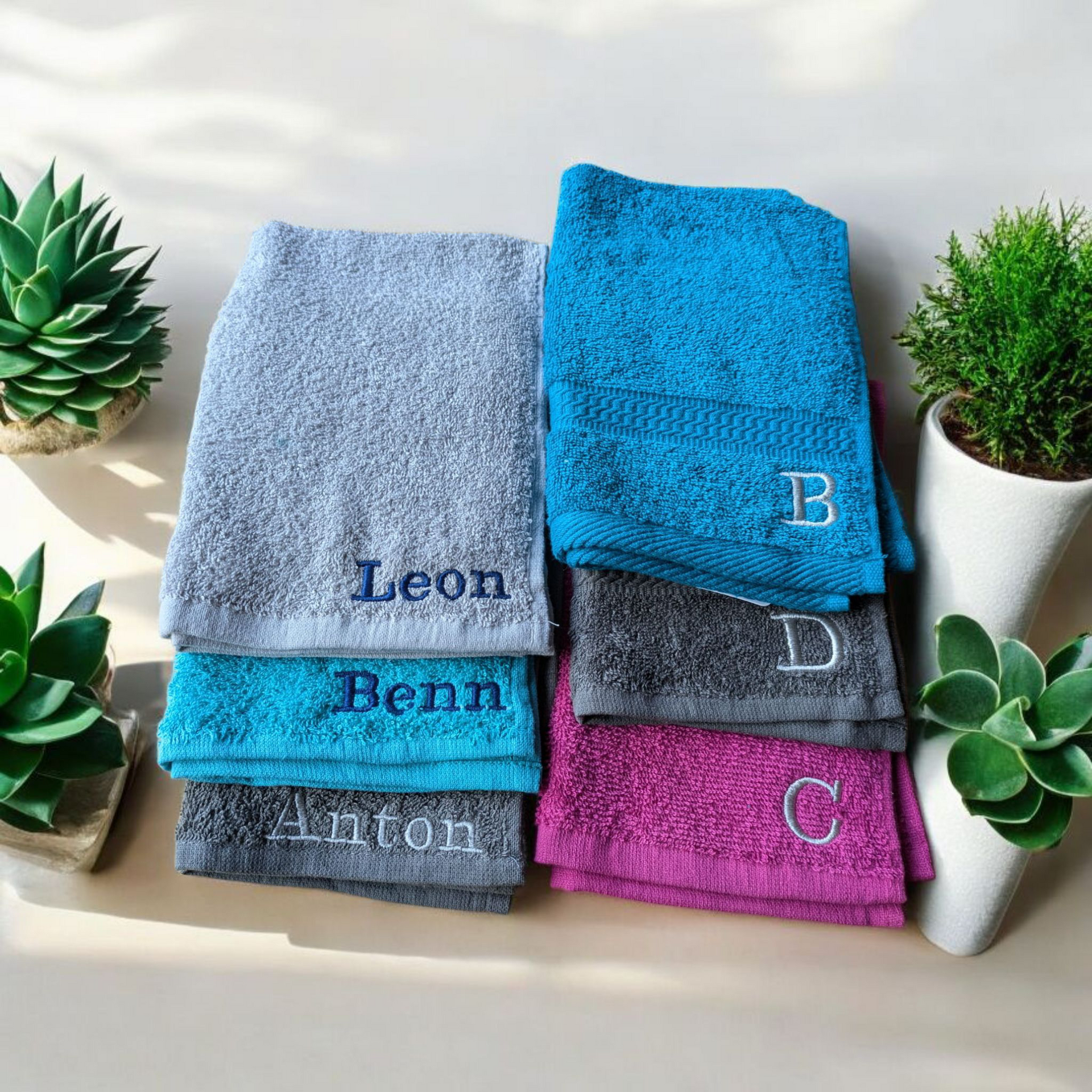 Gestapelte, gefaltete Handtücher: Grau, Türkis und Rosa. Gestickte Namen und Initialen. Sukkulenten und Pflanzen im Hintergrund.