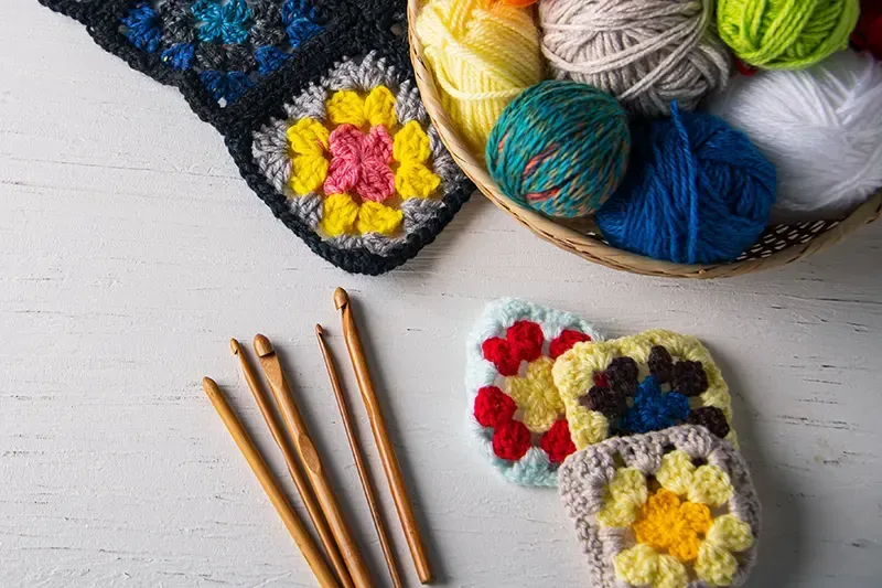 Häkelzubehör: Wollknäuel, Häkelnadeln und Granny Squares auf einer weißen Oberfläche.