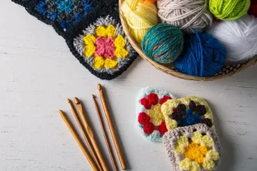 Häkelzubehör: Wollknäuel, Häkelnadeln und Granny Squares auf einer weißen Oberfläche.