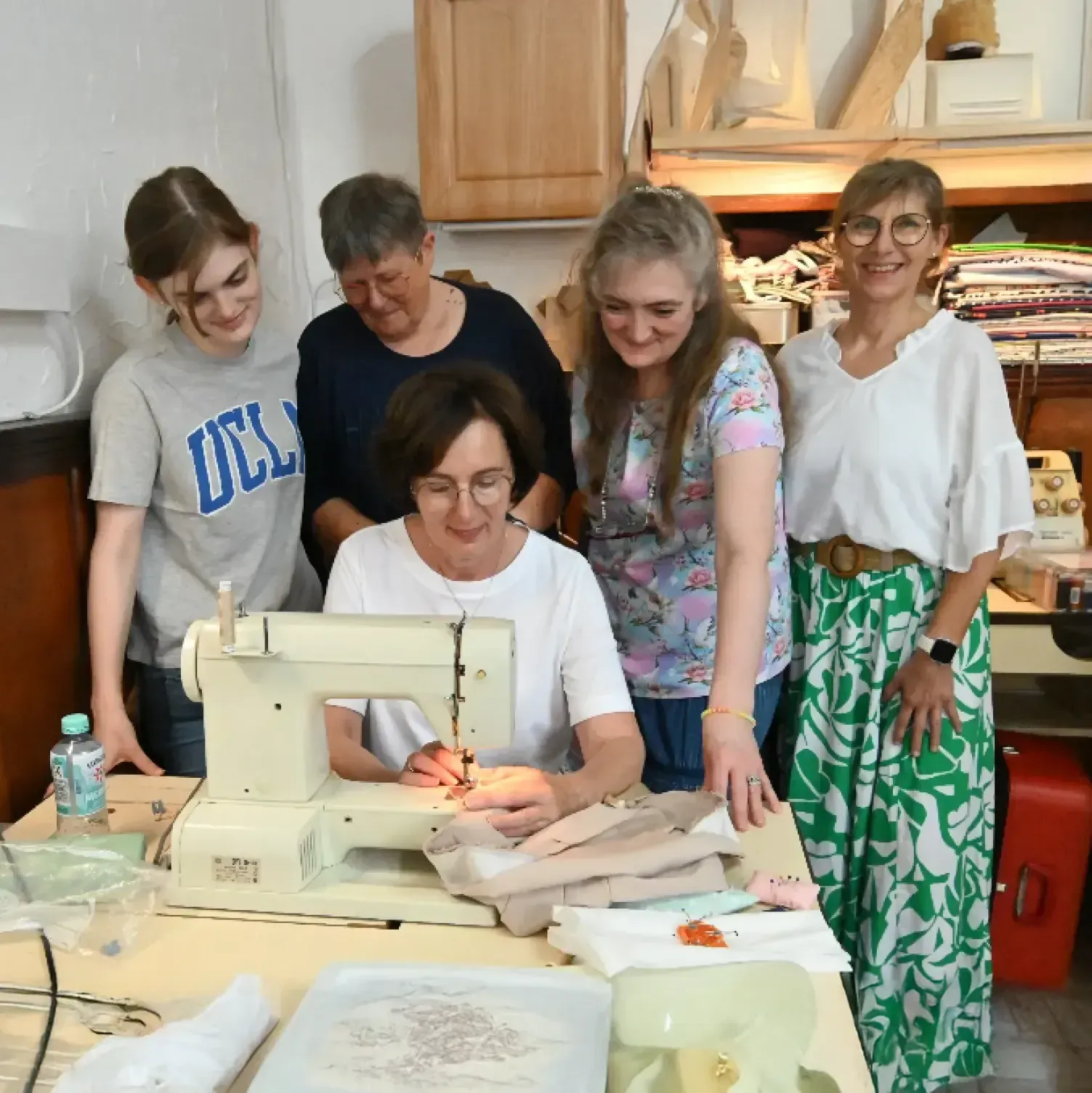 Fünf Frauen versammeln sich um eine Nähmaschine. Eine näht, die anderen schauen zu. Lichtdurchfluteter Raum, im Hintergrund Stoffe.