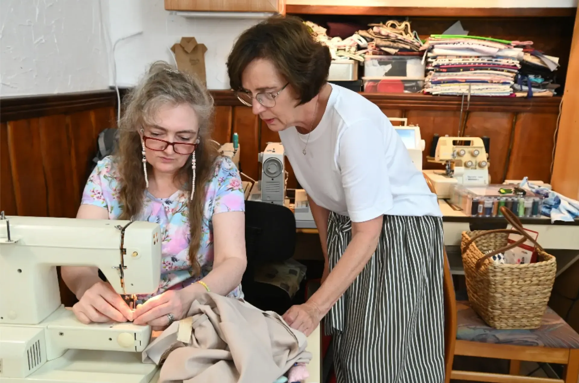 Frau näht an der Maschine, Lehrerin schaut im Nähzimmer mit Stoff und Zubehör zu.