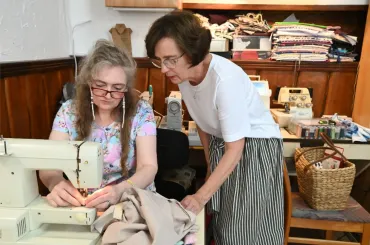 Frau näht an der Maschine, Lehrerin schaut im Nähzimmer mit Stoff und Zubehör zu.