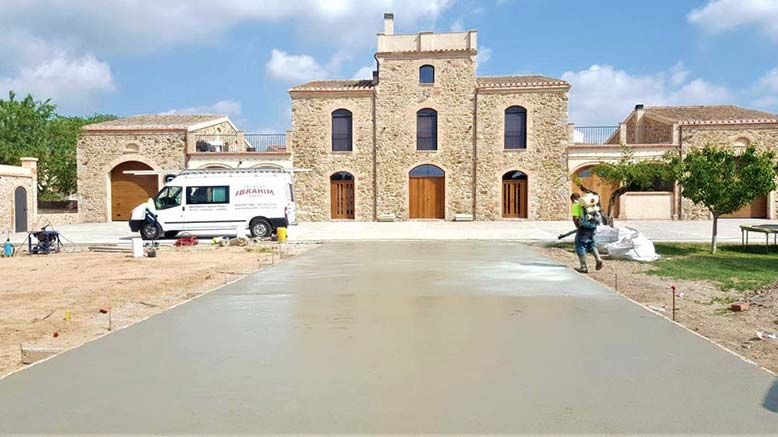 Edificio de piedra con puertas y ventanas arqueadas, una furgoneta blanca aparcada a la izquierda y una persona caminando por una nueva entrada de hormigón.