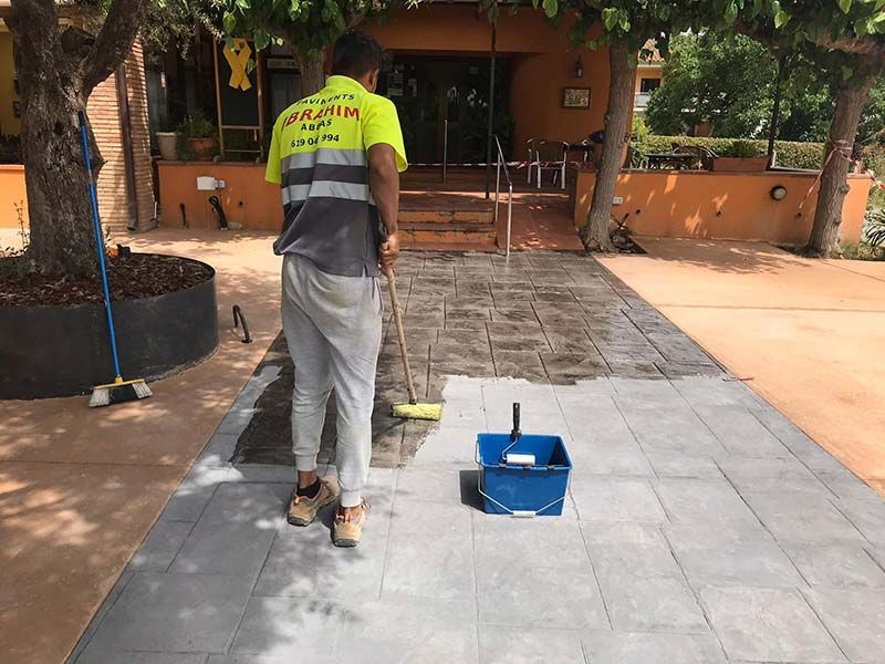 Un trabajador barre un camino de baldosas al aire libre con una escoba y un recogedor cerca de un edificio naranja.