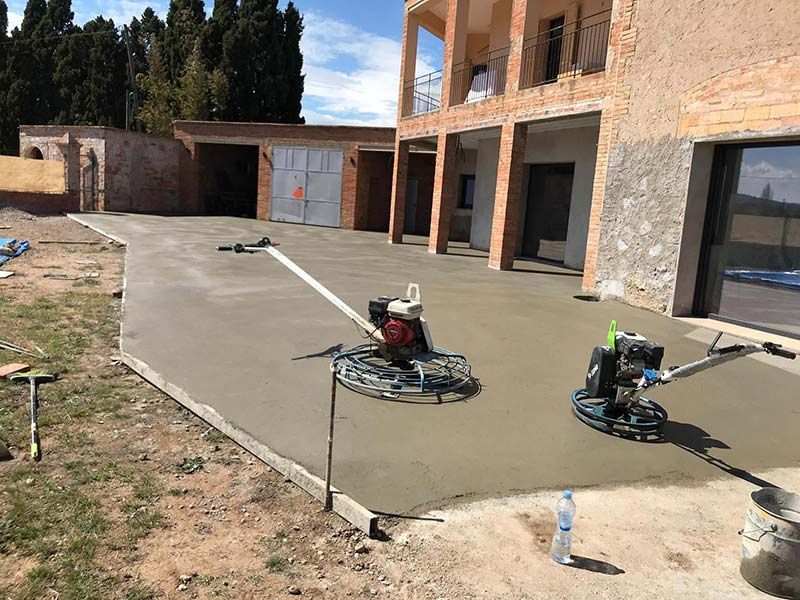Patio de hormigón fresco en construcción junto a una casa de ladrillo, con dos allanadoras mecánicas sobre la losa.