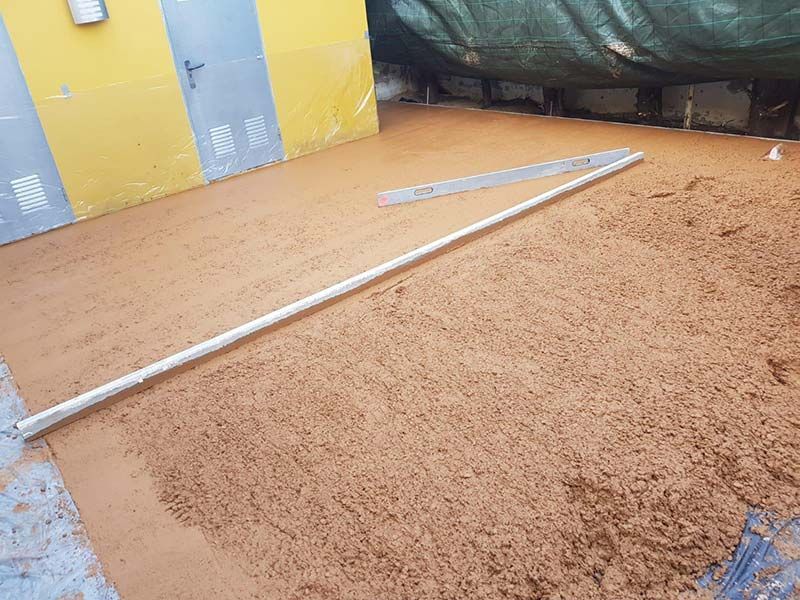 Área arenosa vacía con varillas de metal dispersas junto a una pared amarilla y un fondo de lona oscura.