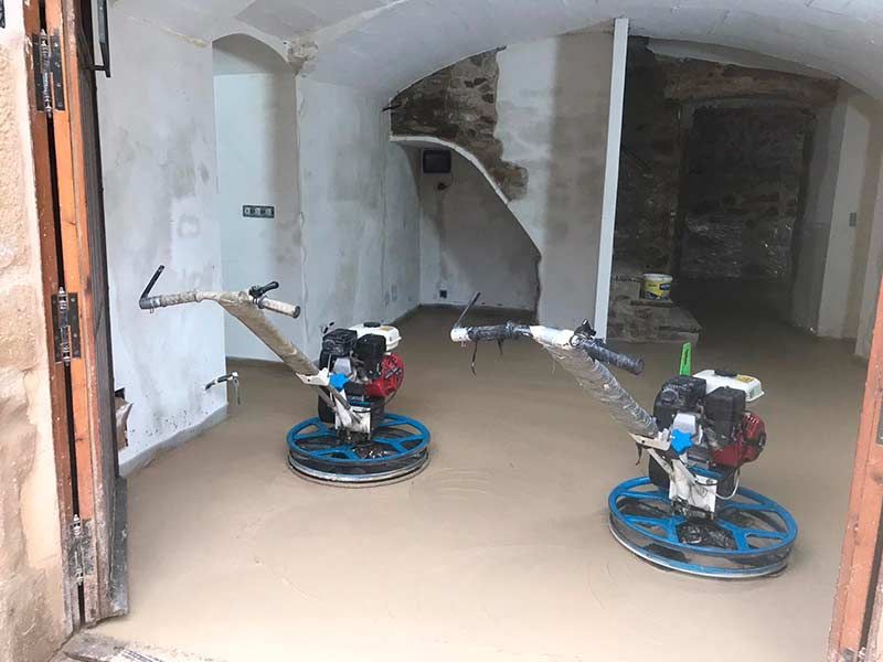 Dos pulidoras de suelos sobre el suelo polvoriento de una habitación sin terminar, con paredes blancas y aberturas arqueadas.