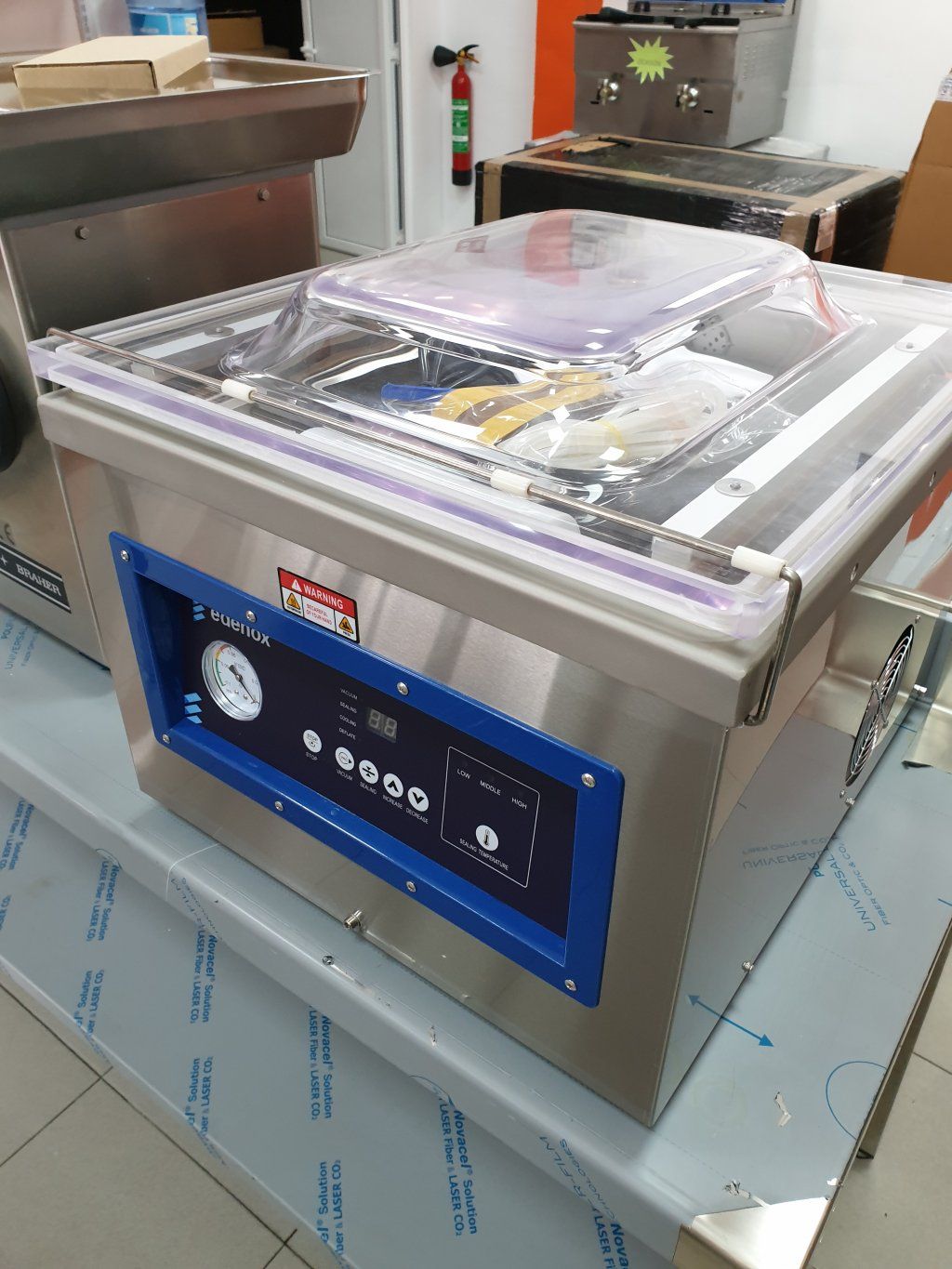 Máquina selladora al vacío de acero inoxidable con panel de control azul y tapa transparente.