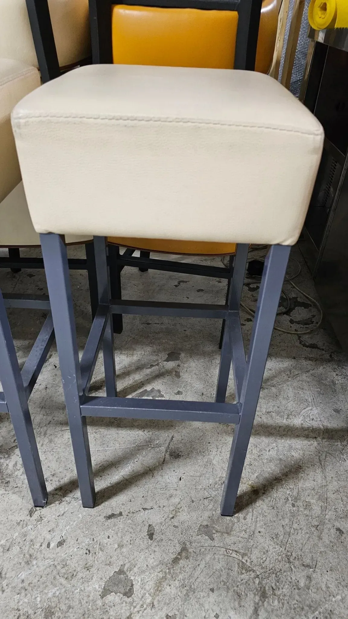Un taburete alto con asiento cuadrado de color marrón claro y patas de metal gris, en una habitación con otros taburetes.