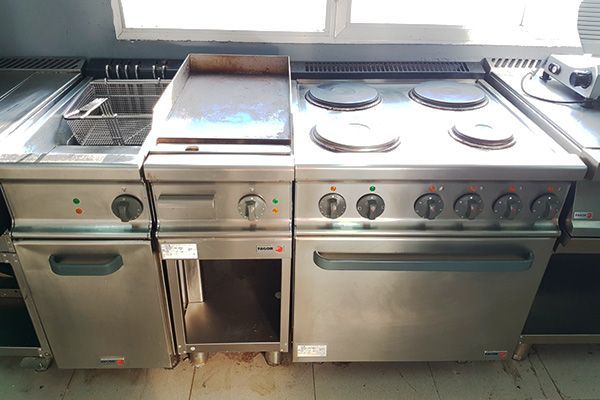 Electrodomésticos de cocina comerciales en acero inoxidable: freidora, plancha, cocina eléctrica con horno, en local luminoso.