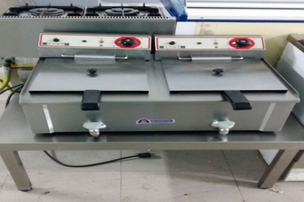 Dos freidoras comerciales plateadas sobre una mesa de acero inoxidable con quemadores de gas al fondo.