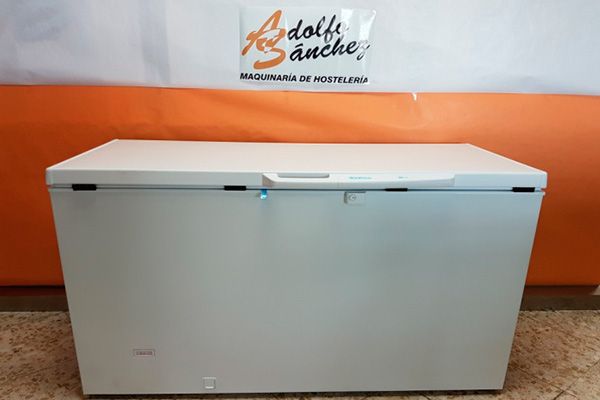 Congelador horizontal de color blanco con asa, sobre un suelo delante de un fondo naranja y blanco con el cartel de una empresa.