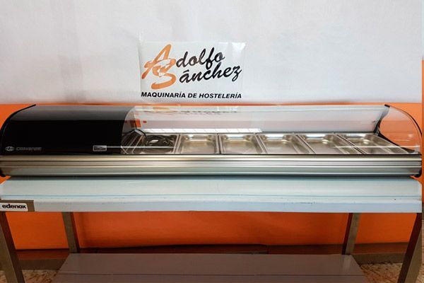 Vitrina refrigerada para alimentos con tapa negra y cubetas de acero inoxidable, sobre un estante de color azul claro, en una tienda.