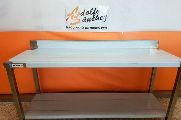 Mesa de trabajo de acero inoxidable de color blanco con estante, situada frente a una pared naranja y blanca.